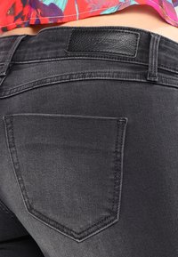 Jeans de mezclilla negra con una textura suave, que cuentan con bolsillos traseros y una etiqueta de marca de cuero. Las costuras visibles complementan el diseño general.