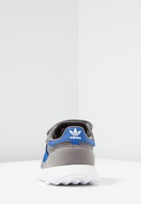 Harmaa urheilukenkä sinisin yksityiskohdin. Kenkässä on teksturoitu mokkanahkainen päällinen, valkoinen kumipohja ja Adidas-logo kannassa. Takakuva.