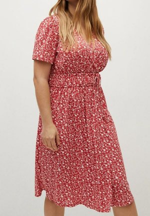 Frau trägt ein rotes Wickelkleid mit kurzen Ärmeln und weißen Blumenmustern, steht vor einem schlichten hellen Hintergrund.