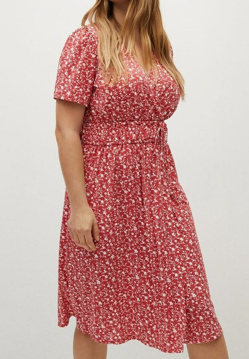 Femme portant une robe portefeuille rouge à manches courtes avec des motifs floraux blancs, debout devant un fond clair uni.