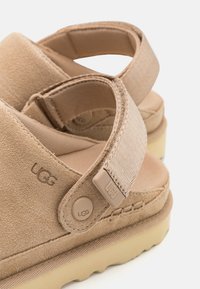 Beige suede sandals with textured straps, featuring a rubber sole and a logo button accent. The design includes a soft, comfortable fit.