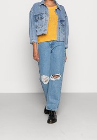 Jeansjacka, gul stickad tröja och rivna ljusblå jeans. Svarta snörkängor. Avslappnad outfit med lös passform och synlig textur.