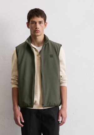 Jeune homme portant une veste sans manches zippée vert olive par-dessus une chemise beige à manches longues et un pantalon noir, debout devant un mur clair uni.