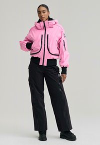 Nő élénk rózsaszín kapucnis dzsekiben, fekete cipzáras részletekkel és fekete terepnadrágban, egyik kezét a zsebében tartva, egyszerű háttér előtt áll.