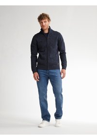 Marineblauer Zip-Pullover mit geripptem Kragen und Bündchen, aus strukturiertem Strick. Kombiniert mit blauen Jeans und weißen Sneakers, vor einem hellen Hintergrund stehend.