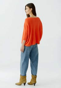 Übergroßer orangefarbener Pullover mit gerippten Ärmeln, kombiniert mit lockeren blauen Jeans und beigen Ankle Boots mit spitzem Absatz. Seitenansicht.