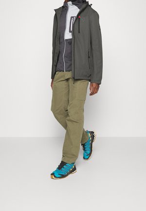 Odporna na warunki pogodowe szara kurtka z kapturem na jasnoszarej warstwie środkowej. Oliwkowozielone spodnie typu cargo i niebieskie buty sportowe z czarnymi akcentami.