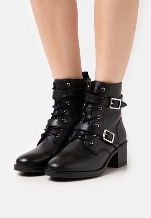 Bottines en cuir noir à lacets, avec deux brides bouclées et talons carrés, portées sur des jambes nues sur fond blanc.