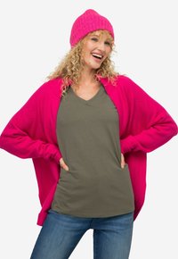 Jasnoróżowy kardigan na oliwkowej zielonej koszulce z dekoltem w serek, zestawiony z dżinsami. Kardigan ma luźny krój i dzianinową strukturę.