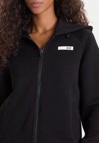 Sudadera negra con cremallera hecha de una tela suave y texturizada, que cuenta con capucha, bolsillos laterales y una pequeña etiqueta con "EA7" en la zona del pecho.