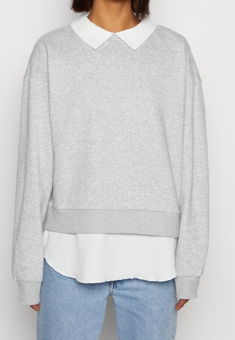 Personne portant un pull gris clair avec une chemise blanche col pointu en dessous et un jean bleu clair, vue des épaules aux hanches.