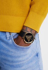 Montre noire avec accents dorés, présentant un motif triangulaire sur le cadran. Portée avec un pull en maille jaune et un jean bleu clair.