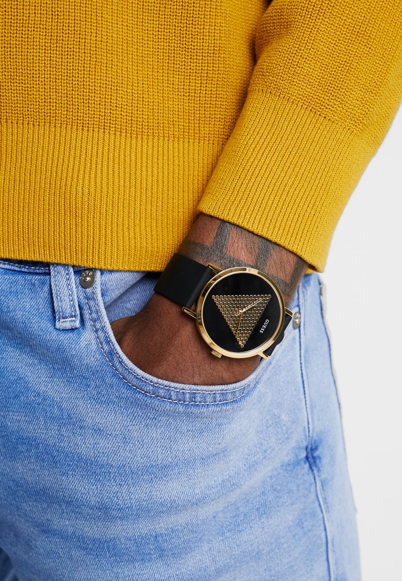 Montre noire avec accents dorés, présentant un motif triangulaire sur le cadran. Portée avec un pull en maille jaune et un jean bleu clair.