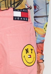 Pantalones de mezclilla rosa con una gran cara sonriente amarilla con el texto "NY" y una pequeña etiqueta de Tommy Jeans. Tejido texturizado con detalles de costura en contraste.