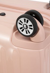 Valigia rigida rosa con superficie liscia, dotata di ruote in gomma nera e dettagli argentati in un design radiale. Chiusura con zip visibile.
