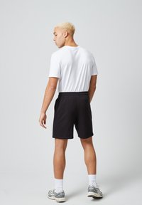 Zwarte sportshorts met een elastische tailleband, gecombineerd met een eenvoudig wit T-shirt. Het model is van achteren te zien, rechtopstaand.
