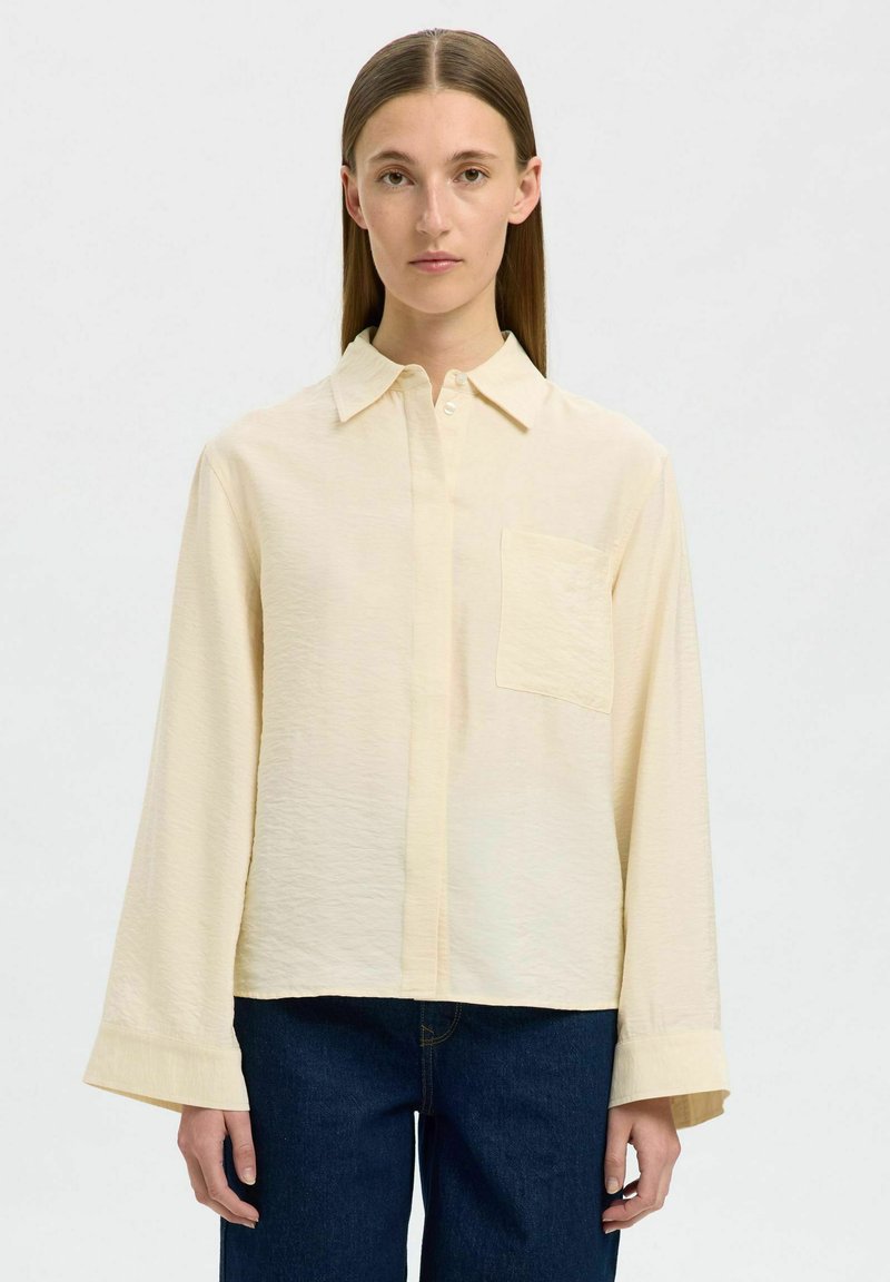 Camisa de manga larga amarilla clara con cuello abotonado, un único bolsillo en el pecho y una tela texturizada. Llevada con jeans azul oscuro.