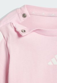 Camiseta de algodón rosa claro con botones de presión en el escote. Cuenta con un cuello acanalado y un logotipo blanco impreso en el frente.