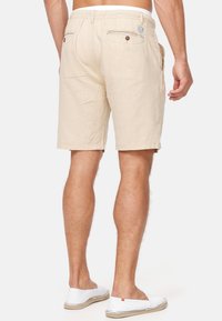 Man i beiga shorts med knäppta bakfickor och vita slip-on skor, stående mot en enfärgad vit bakgrund.