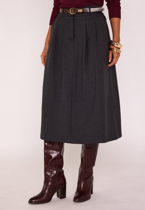 Femme portant une jupe midi plissée gris foncé avec une ceinture fine, un haut à manches longues bordeaux, des bracelets dorés et des bottes montantes bordeaux brillantes.