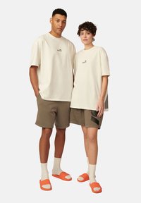 Cremefarbene T-Shirts, olivgrüne Shorts und orangefarbene Sandalen an zwei Modellen. Die Shorts haben ein einzigartiges Designelement. Strukturierter Stoff ist sichtbar.