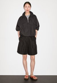 Pullover nero a mezzo zip con colletto alto, abbinato a pantaloni corti neri larghi e infradito marroni con cinturini rossi, su uno sfondo chiaro.
