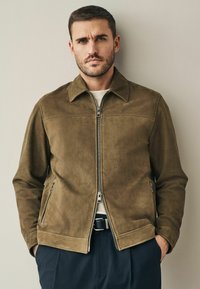 Bruine suède jas met een spitse kraag, ritssluiting aan de voorkant, twee zijzakken en een gladde textuur, gedragen over een licht shirt en donkere broek.