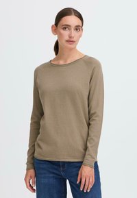 Langermet topp med rund hals i lys brun stoff med teksturert overflate. Har ribbede mansjetter og en avslappet passform, kombinert med blå jeans.