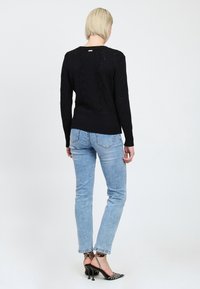 Donna con capelli biondi corti che indossa un maglione nero a fantasia, jeans azzurri chiari e sandali neri con tacco alto e cinturini, in piedi di spalle.