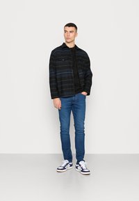 Jovem em pé contra um fundo neutro, vestindo uma jaqueta listrada em preto e azul, camisa preta, jeans azuis e tênis brancos.