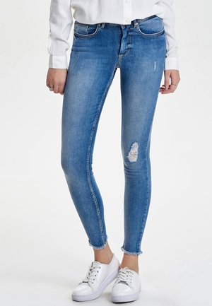 Kvinde iført lyseblå revnede skinny jeans med frynset kant, hvid knappet skjorte og hvide sneakers, stående mod en ensfarvet baggrund.