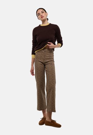 Un pull marron avec des accents jaunes, associé à un pantalon beige court ornés d'un imprimé léopard noir. Le modèle porte des chaussures en daim marron.