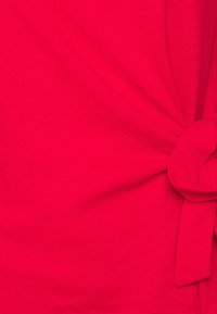 Camiseta roja de algodón suave, con un detalle anudado en el costado para añadir interés de diseño. Textura suave y forma ajustada.