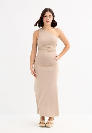 Vestido largo beige de un solo hombro con cuerpo fruncido. Tela suave y elástica con silueta ajustada. Combinado con sandalias de plataforma beige.