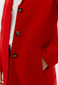 Red wool coat featuring a soft texture, five buttons in dark shades, and a side pocket. The design has a tailored fit and classic collar.