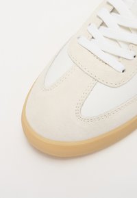Beige suede sneaker with white leather accents, rounded toe, and rubber gum sole. Features white laces and stitched detailing.