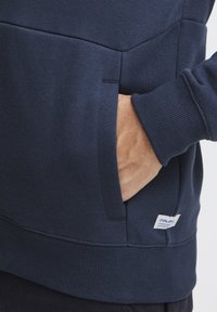 Marineblå hettegenser med kengurulomme, ribbede mansjetter og nederste kant, samt en liten logotagg nær bunnsømmen. Strukturert stoff.