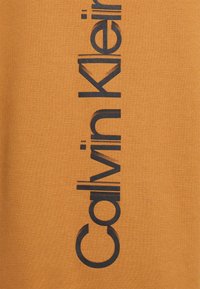Vertikal svart "Calvin Klein" logotext på en senapsgul tygbakgrund.