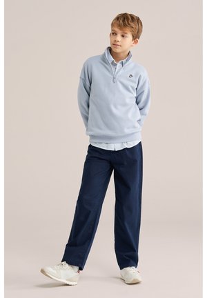 Dječak stoji s rukama iza leđa, nosi svijetloplavu majicu sa zatvaračem do pola, prugastu košulju, mornarsko plave hlače i bijele tenisice na bež pozadini.