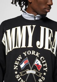 Felpa nera con grande testo bianco e oro "TOMMY JEAN" e un logo circolare con bandiere doppie e banner, tessuto testurizzato con colletto zip.