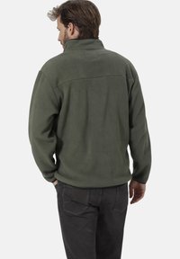 Olivgrüne Fleece-Jacke mit hohem Kragen, texturierter Oberfläche und rechteckigem Rückenpatch. Kombiniert mit dunklen Jeans.