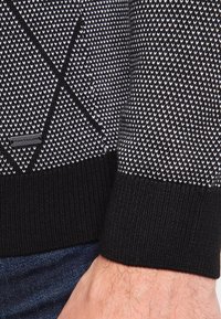 Musta-valkoinen kuviollinen neulepaita, jossa on teksturoitu kangas, yhtenäinen musta reuna ja logoplästi alareunan vasemmassa kulmassa.
