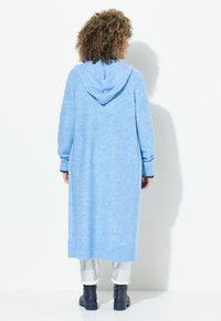 Blå lång cardigan med huva, ribbade ärmslut och nederkant, tillverkad av ett mjukt, texturerat tyg. Buren över ljusa byxor och svarta stövlar.