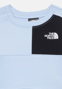 Lys blå genser med svart panel, med rund hals, stikkede detaljer og "The North Face"-logo i hvitt på brystet.