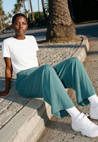 Young woman wearing a white t-shirt, teal wide-leg pants, white socks, and white sneakers sitting on a curb near a palm tree on a sunny street.
