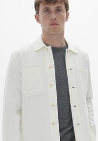 Hvid skjorte med klassisk krave, der har en brystlomme og lyse træknapper, båret over en grå sweater. Glat stofstruktur.