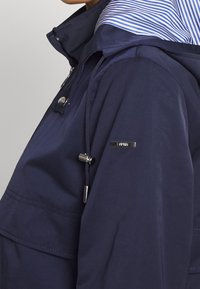 Wodoodporna granatowa kurtka z kapturem. Posiada paskowane podszycie, regulowane sznurki oraz metalowe detale na klatce piersiowej i rękawie.