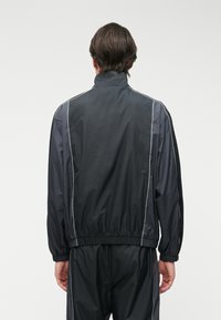 Μαύρο μπουφάν με ψηλό γιακά, ελαστική μέση και λευκές πλαϊνές ρίγες, κατασκευασμένο από ελαφρύ υλικό.