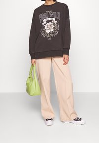 Tmavě šedá oversized mikina s květinovým grafickým designem, kombinovaná s béžovými kalhotami širokého střihu a zelenou kabelkou. Černobílé tenisky dokončují vzhled.