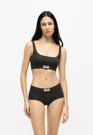 Femme aux longs cheveux foncés portant un soutien-gorge de sport noir et un slip taille haute assorti avec des étiquettes de marque sur la poitrine et la taille.